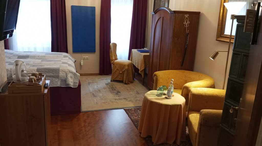 Doppelzimmer mit Bett und grauer Tagesdecke, rotem Vorhang und blauem Wandpaneel, Schreibtisch mit Stuhl, Holzschrank, gelbem Sessel und rundem Tisch mit Wasserflasche und Gläsern.