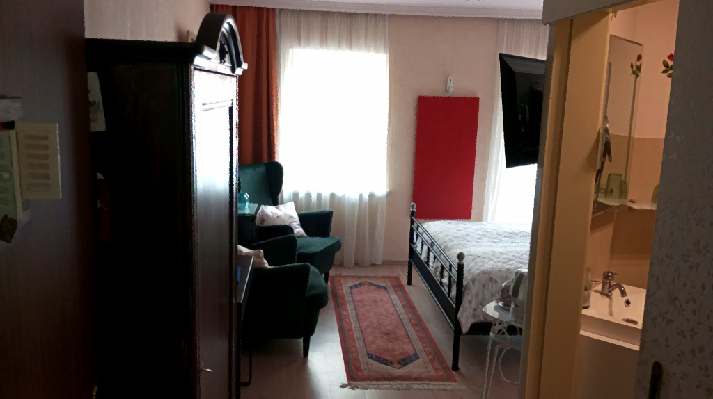 Doppelzimmers „English Rose“ mit Metallbett, grünem Sessel, rotem Infrarotpanel und Blick durch den Türrahmen ins Bad.
